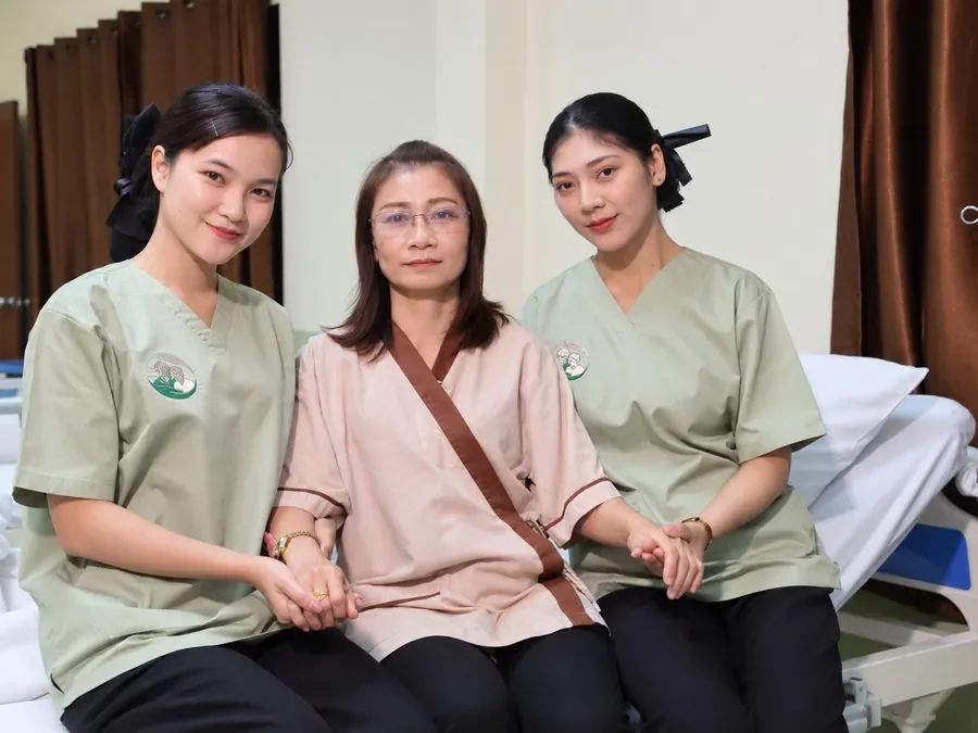 ดูแลด้วยความอบอุ่นเหมือนคนในครอบครัว | Omkod Nursing Home Rama 3 ผู้ดูแล 2 คนนั่งจับมือพูดคุยกับผู้พักบนเตียงในห้องพัก อ้อมกอด โฮมแคร์ภิบาล สาขาพระราม 3