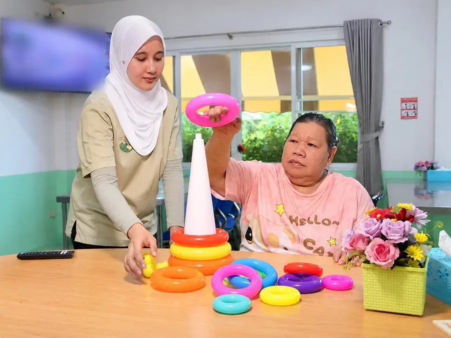 คุณยายหยิบห่วงสวมหลัก ผู้ดูแลช่วยจัดห่วงในห้องกิจกรรม