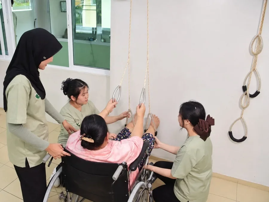 คุณยายนั่งรถเข็นดึงห่วงรอกบริหารแขน มีผู้ดูแลประคองอยู่ข้าง ๆ