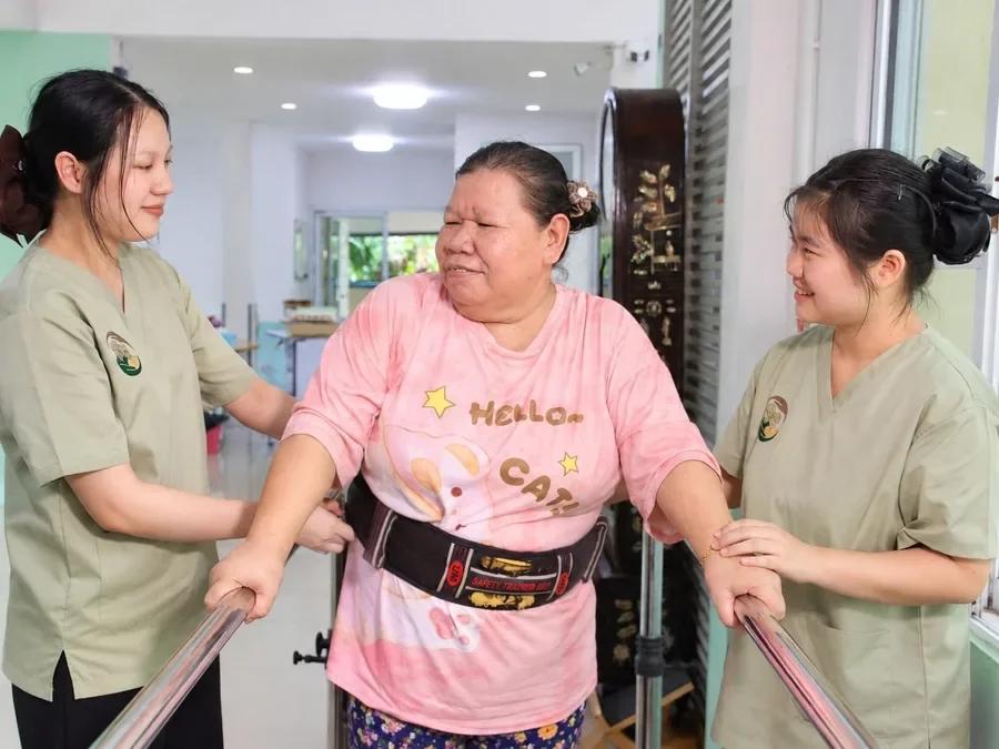 ผู้ดูแลประคองคุณยายเดินในราวจับ สวมเข็มขัดพยุงตัวเพื่อความปลอดภัย