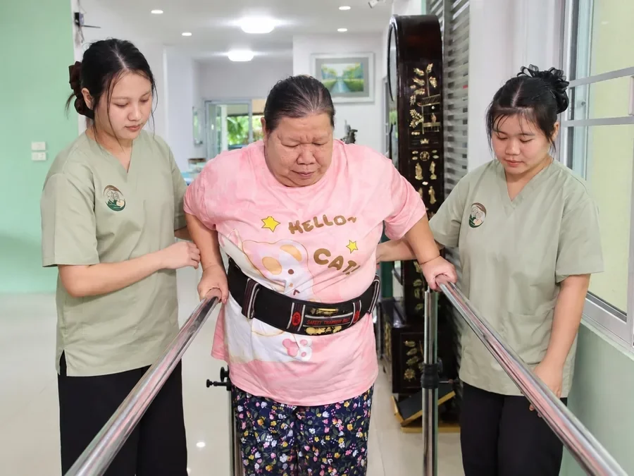 คุณยายฝึกยืนเดินในราวจับ สวมเข็มขัดพยุงตัว มีผู้ดูแลประกบสองข้าง